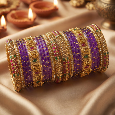 A stack of ornate gold-plated bangles with purple, red, and green stone accents, displayed on draped satin fabric with blurred diya candles in the background.