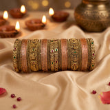 A set of traditional Indian bangles (batches) featuring gold-plated filigree and pink stone-encrusted bands with small red and green stone accents, displayed on draped beige satin fabric with lit diya candles in the background.