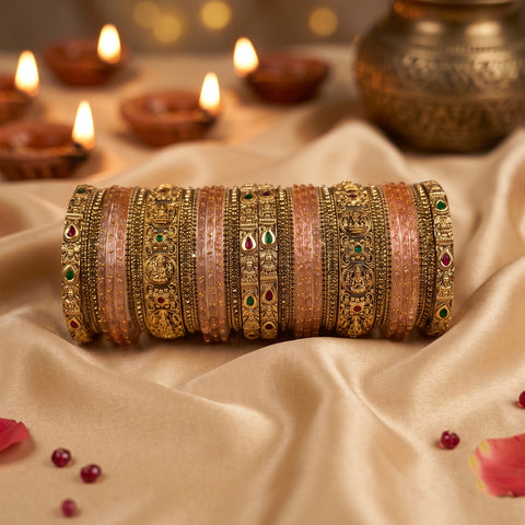 A set of traditional Indian bangles (batches) featuring gold-plated filigree and pink stone-encrusted bands with small red and green stone accents, displayed on draped beige satin fabric with lit diya candles in the background.