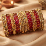 A stack of traditional Indian bangles, featuring alternating rows of red stone-covered bangles and ornate gold-plated bangles with deity motifs and small green and red gemstone accents, displayed on draped beige fabric.