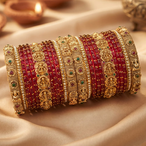 A stack of traditional Indian bangles, featuring alternating rows of red stone-covered bangles and ornate gold-plated bangles with deity motifs and small green and red gemstone accents, displayed on draped beige fabric.