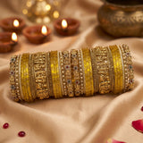 A stack of ornate gold and yellow bangles with intricate stone and metallic detailing, arranged on draped beige satin fabric with lit diya candles and a brass pot in the softly blurred background.