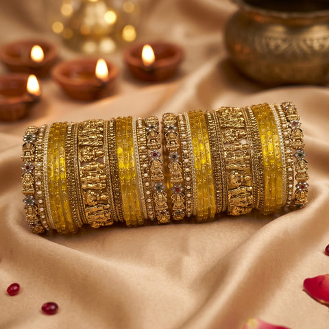 A stack of ornate gold and yellow bangles with intricate stone and metallic detailing, arranged on draped beige satin fabric with lit diya candles and a brass pot in the softly blurred background.