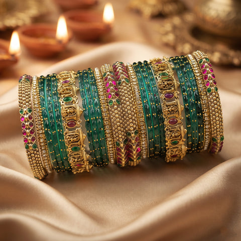 A stack of ornate Indian bangles (batches) made of gold-plated metal, decorated with green stone-encrusted bands and pink stone accents, resting on draped beige satin fabric with blurred diya candles in the background.