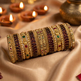 A stack of ornate gold-toned bangles with red, green, and dark maroon stone accents, displayed on draped satin fabric with lit diya candles in the background.