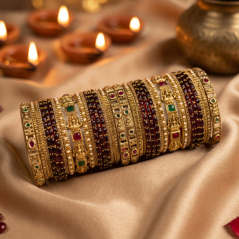 A stack of ornate gold-toned bangles with red, green, and dark maroon stone accents, displayed on draped satin fabric with lit diya candles in the background.