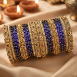 A stack of traditional Indian bangles (kara) featuring royal blue beaded bands, ornate gold-plated filigree with deity motifs, and small green and red stone accents, displayed on draped beige satin fabric with blurred diya candles in the background.