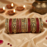 A stack of traditional Indian bangles (batches) featuring red beaded bracelets alongside gold-plated bangles with intricate filigree, green and red stone accents, displayed on a beige satin fabric with lit diya candles in the background.