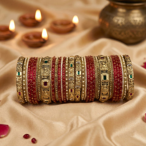 A stack of traditional Indian bangles (batches) featuring red beaded bracelets alongside gold-plated bangles with intricate filigree, green and red stone accents, displayed on a beige satin fabric with lit diya candles in the background.
