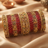 A stack of traditional Indian bridal bangles, featuring red stone-encrusted bands alongside ornate gold-plated filigree bangles with green and red gemstone accents, displayed on draped beige satin fabric.