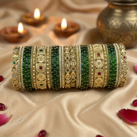A set of ornate Indian bangles (kara) displayed on a beige satin fabric. The bangles feature intricate gold-plated filigree, rows of green beads, and small red and green stone accents.