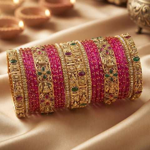 A stack of ornate Indian bangles (kara) featuring bright pink stone-set bands alongside detailed gold-plated filigree bangles with red and green stone accents, displayed on a beige satin fabric with blurred diya lights in the background.