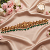 An ornate gold-toned traditional necklace with intricate filigree work, red stone accents, and dangling green beads, arranged on draped beige satin fabric with pink roses and a small lit candle.