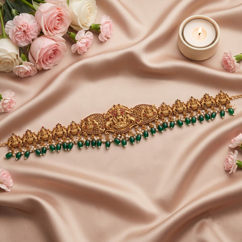 An ornate gold-toned traditional necklace with intricate filigree work, red stone accents, and dangling green beads, arranged on draped beige satin fabric with pink roses and a small lit candle.