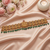 An ornate gold-toned traditional necklace with detailed filigree, red and green stone accents, and a row of dangling green beads, arranged on draped blush satin fabric with pink flowers and a lit candle.