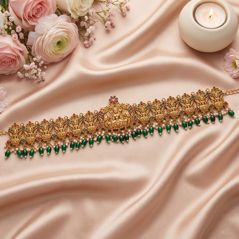 An ornate gold-toned traditional necklace with detailed filigree, red and green stone accents, and a row of dangling green beads, arranged on draped blush satin fabric with pink flowers and a lit candle.