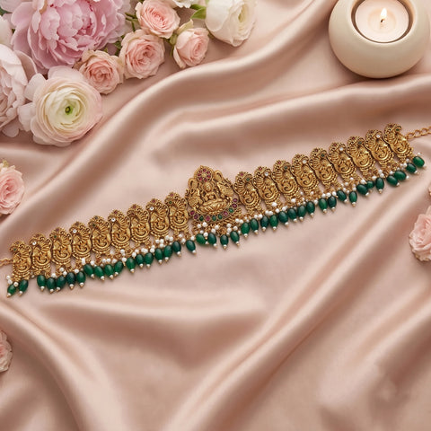 An ornate gold-toned traditional necklace with a central deity motif, decorated with rows of small white pearls and dangling green beads, displayed on draped blush satin fabric with pink flowers and a lit candle.