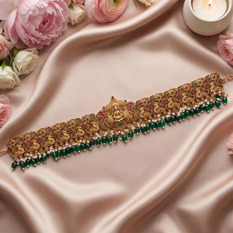 An ornate gold-toned traditional necklace with intricate filigree, small pink gem accents, and dangling green beads and white pearls, arranged on draped blush satin with flowers and a lit candle around it.