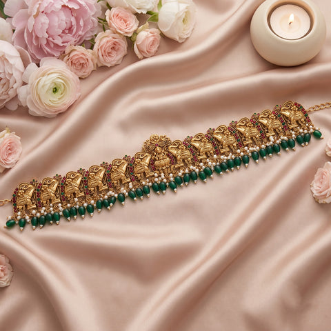 A gold-toned traditional Indian choker necklace with elephant motifs, green bead drops, and small pearl-like accents, arranged on draped blush satin fabric with pink and white roses and a lit candle.