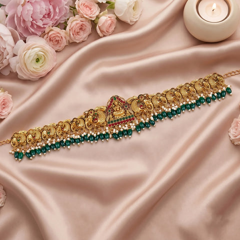 An ornate gold-toned traditional necklace with elephant motifs, red and green stone accents, and a row of dark green beads, styled on draped blush satin fabric with pink flowers and a lit candle.