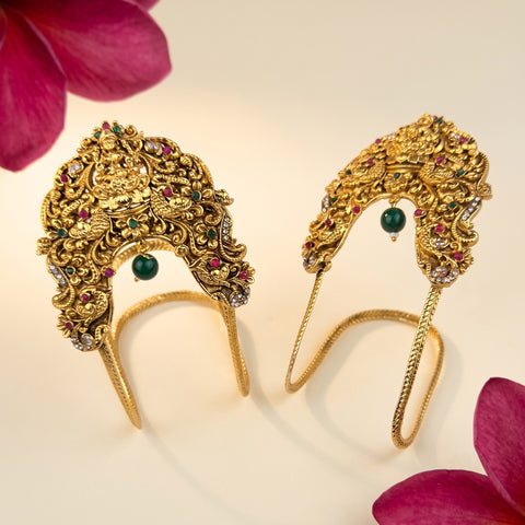 A pair of ornate gold headbands decorated with intricate filigree, small green and red gemstones, and a central green bead, displayed on a light cream background with pink flower petals in the corners.