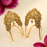 A pair of ornate gold-toned traditional Indian headpieces with intricate filigree, small hanging gold bells, and colorful gemstone accents, displayed on a light cream background with pink flower petals in the corners.