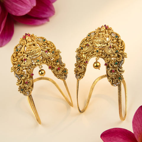 A pair of ornate gold-toned traditional Indian headpieces with intricate filigree, small hanging gold bells, and colorful gemstone accents, displayed on a light cream background with pink flower petals in the corners.