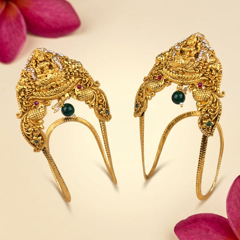 A pair of ornate gold-toned traditional hair ornaments with intricate filigree, small green and red gemstone accents, and curved lower bands, displayed on a light cream background with pink flower petals at the corners.