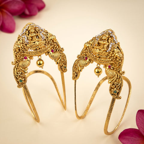 A pair of ornate gold-toned traditional Indian hair ornaments (Jhumkas) with detailed filigree, small hanging gold beads, and tiny colored gem accents, displayed on a light cream background with decorative pink flower petals in the corners.