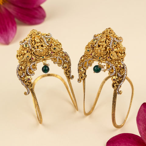 A pair of ornate gold filigree headbands decorated with rhinestones and green bead accents, displayed on a light beige background with pink flower petals.