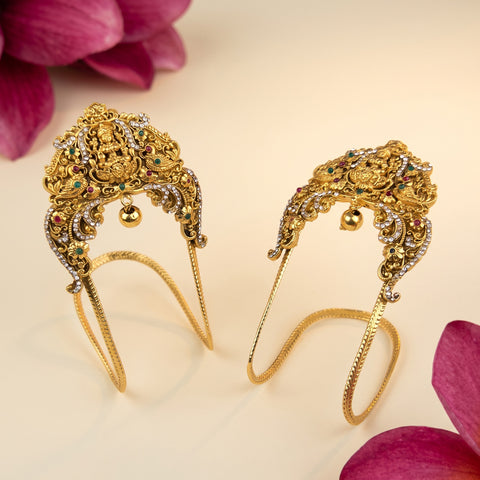 A pair of ornate gold filigree headbands decorated with small green and clear stones, displayed on a light beige background with pink flower petals in the corners.