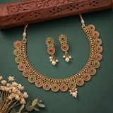 An ornate gold-toned Indian bridal necklace and matching drop earrings set, decorated with green and red stones and white pearl accents, displayed on a dark green background with dried flowers and a carved wooden prop.