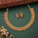 Gold-toned traditional necklace and matching earrings with red and green stone accents, arranged on a dark green background with dried flowers and a carved wooden prop.