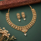 Gold-toned traditional necklace and matching earrings with pearl-like white stones and small red and green stone accents, arranged on a dark green background with decorative dried flowers and a carved wooden prop.