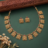 Gold-toned traditional necklace and matching stud earrings with ornate square motifs and small red oval stones, arranged on a dark green surface with dried flowers and a carved wooden prop.