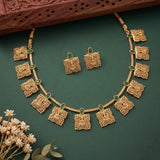 Gold-toned traditional necklace and matching earrings with ornate square motifs and small green stone accents, arranged on a dark green background with decorative dried flowers and a carved wooden prop.
