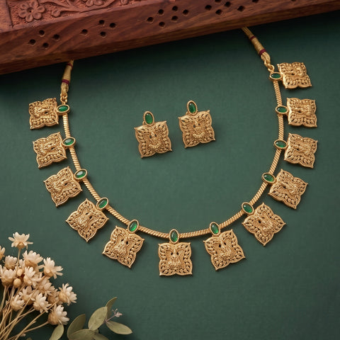 Gold-toned traditional necklace and matching earrings with ornate square motifs and small green stone accents, arranged on a dark green background with decorative dried flowers and a carved wooden prop.