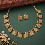 Gold-toned traditional jewelry set featuring a necklace and matching earrings with ornate square motifs and small multicolored stone accents, arranged on a dark green background with decorative dried flowers and a carved wooden prop.