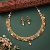Gold-plated Indian style necklace and matching earrings set with red, green, and white stones, displayed on a dark green background with a carved wooden prop and dried flowers.