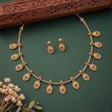 Gold-toned traditional necklace and matching drop earrings with small red stone accents, arranged on a dark green background with decorative dried flowers and a carved wooden prop.