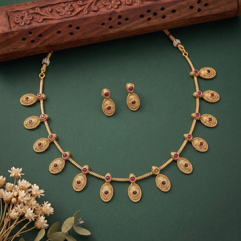 Gold-toned traditional necklace and matching drop earrings with small red stone accents, arranged on a dark green background with decorative dried flowers and a carved wooden prop.