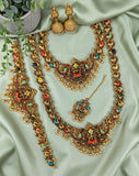 An ornate gold-toned Indian jewelry set laid out on a light sage green satin fabric, including a statement necklace, matching bracelet, earrings, and a pendant with colorful enamel details.