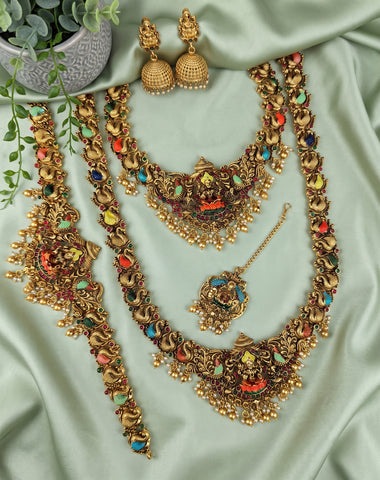 An ornate gold-toned Indian jewelry set laid out on a light sage green satin fabric, including a statement necklace, matching bracelet, earrings, and a pendant with colorful enamel details.