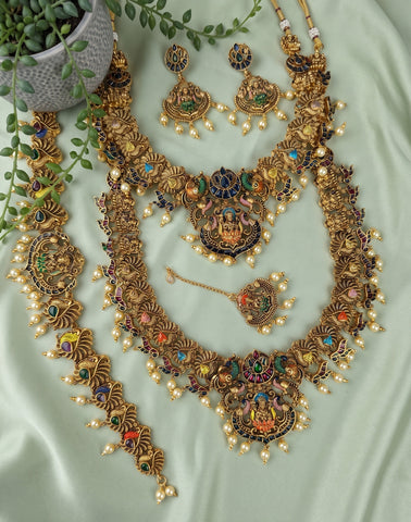 An ornate traditional Indian jewelry set laid out on a light sage green fabric background, featuring a gold-plated necklace, matching earrings, bracelet, and pendant with colorful enamel details and small pearl-like beads. A small potted plant is visible in the top-left corner.