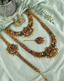 An ornate gold-toned jewelry set arranged on pale mint green satin fabric, including a necklace, matching earrings, a bracelet, and a small pendant, decorated with multicolored stones and pearl-like beads. Greenery and a small pot are placed as styling props.