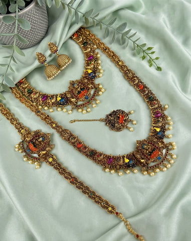 An ornate gold-toned jewelry set arranged on pale mint green satin fabric, including a necklace, matching earrings, a bracelet, and a small pendant, decorated with multicolored stones and pearl-like beads. Greenery and a small pot are placed as styling props.