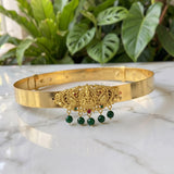 A gold-plated traditional Indian choker necklace with a detailed Ganesha ornament and hanging green bead tassels, displayed on a white marble surface with green plants in the background.