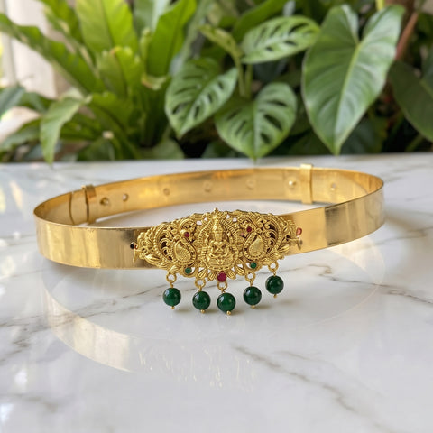 A gold-plated traditional Indian choker necklace with a detailed Ganesha ornament and hanging green bead tassels, displayed on a white marble surface with green plants in the background.