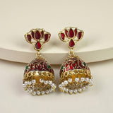 A pair of traditional Indian gold-plated jhumka earrings with red enamel, green stone accents, and dangling white pearl chains, displayed on a light beige background.