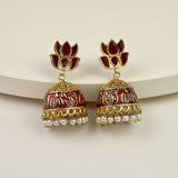 A pair of gold-plated Indian jhumka earrings with deep red floral studs and red-and-gold patterned bell-shaped drops, finished with small white pearl tassels, displayed on a curved cream-colored surface.